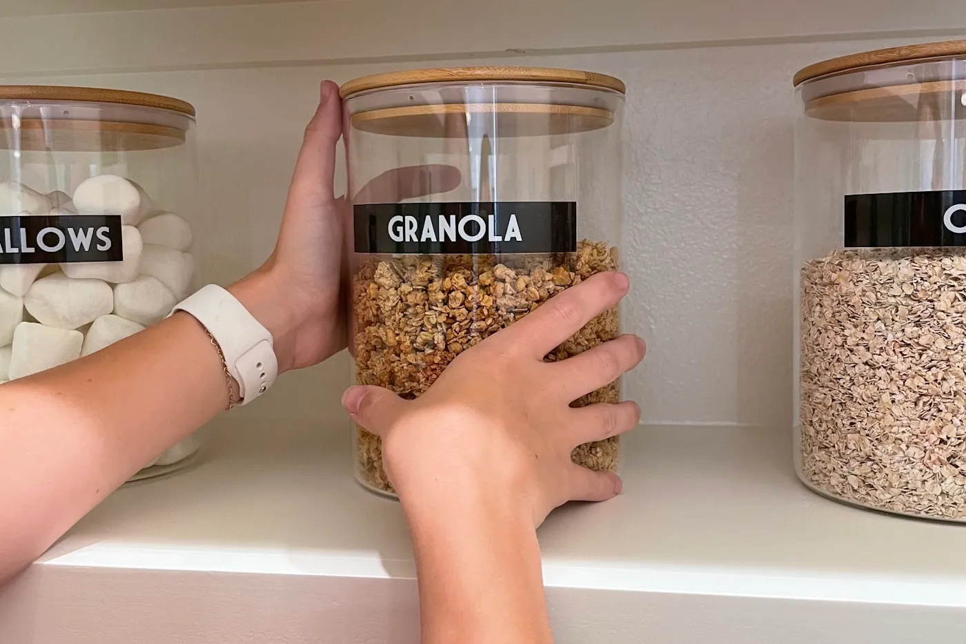 A hand lifting a glass jar of granola from a pantry shelf